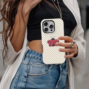 May include: A phone case with a cream-colored background and small black polka dots. A red and black ladybug design is centered on the case. The phone case is being held by a person.