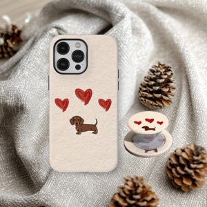Könnte beinhalten: Beigefarbene Handyhülle mit einer Dackel-Illustration und drei roten Herzen. Ein passender Handyhalter mit dem gleichen Design ist ebenfalls zu sehen. Die Artikel befinden sich auf einer strukturierten, hellgrauen Oberfläche mit Tannenzapfen.