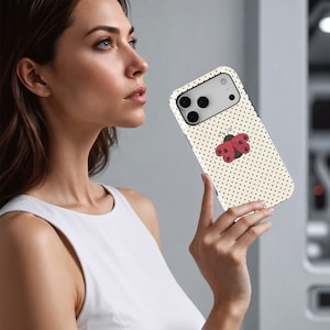 May include: A smartphone case with a cream-colored background and small black polka dots. A red and black ladybug design is centered on the case. The case has a black border and a camera cutout.
