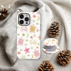 May include: White iPhone case and matching phone grip featuring a watercolor design of sea turtles, crabs, seahorses, starfish, and flowers. The case and grip have a white background.