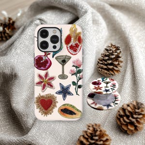 May include: A light pink iPhone case with embroidered designs of a pomegranate, martini glass, star, and other botanical and fruit motifs. A matching pop socket is also visible. The case and pop socket are on a gray fabric background.