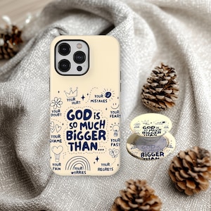 May include: A cream-colored phone case and matching pop socket with the phrase "GOD IS SO MUCH BIGGER THAN..." in navy blue. The case features doodles of a crown, heart, and rainbow. Two pinecones rest on a gray fabric.