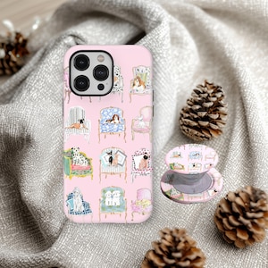 Puede incluir: Funda rosa para iPhone y soporte retráctil para teléfono a juego con un patrón repetido de perros en sillas. La funda tiene un borde negro para la lente de la cámara. Dos piñas se encuentran sobre un fondo de tela gris.