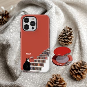 May include: A red phone case and pop socket with a design of a black and white cat, a wine glass, and a staircase of books. The case has the word "Miau!!" printed on it.