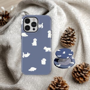 Könnte beinhalten: Eine blaue Handyhülle und ein passender Handyhalter mit einem sich wiederholenden Muster aus weißen Pudel-Illustrationen. Die Hülle liegt auf einer grauen Oberfläche mit Tannenzapfen.