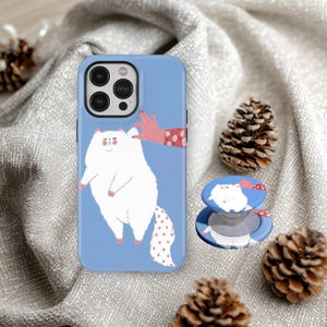 May include: A light blue phone case and matching pop socket featuring a whimsical white cat illustration with a pink hand. The cat has a pink nose and paws, and a dotted tail. The pop socket is round and has a mushroom design.