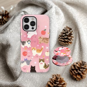May include: Pink iPhone case with a cat pattern, featuring various cat illustrations, apples, and stars. A matching phone grip is also visible. The case and grip are on a gray fabric background, with pinecones.