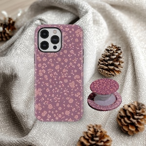May include: A mauve phone case and matching phone grip featuring a delicate floral pattern in shades of pink. The phone case is shown with a phone, and the phone grip is next to it. The background includes pine cones and a gray fabric.