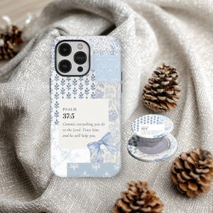 Può includere: Una custodia per telefono e un supporto abbinato blu chiaro e bianco con un design floreale e patchwork. La custodia e il supporto presentano il testo "SALMO 37:5" e una citazione. Le pigne decorative sono sullo sfondo.
