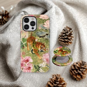 May include: A phone case and phone grip featuring a tiger design. The case and grip have a green background with pink flowers and a tiger illustration. The phone case is on a gray cloth with pine cones.