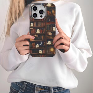 Puede incluir: Una funda de tel&eacute;fono con una ilustraci&oacute;n caprichosa de una estanter&iacute;a llena de libros y peque&ntilde;os fantasmas amigables. El dise&ntilde;o incluye una luna creciente, una linterna y un cielo estrellado, creando una est&eacute;tica acogedora y m&aacute;gica. La funda es sostenida por una persona.