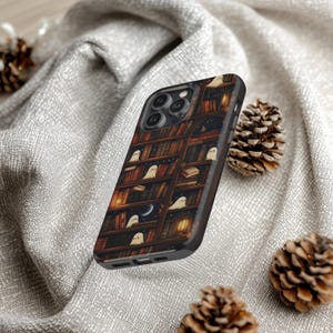 Puede incluir: Una funda negra para smartphone con un dise&ntilde;o caprichoso de una estanter&iacute;a llena de libros y peque&ntilde;os fantasmas amigables. La funda tiene un borde de imitaci&oacute;n de madera oscura y un cielo nocturno con una luna creciente. El tema general es espeluznante y lindo.