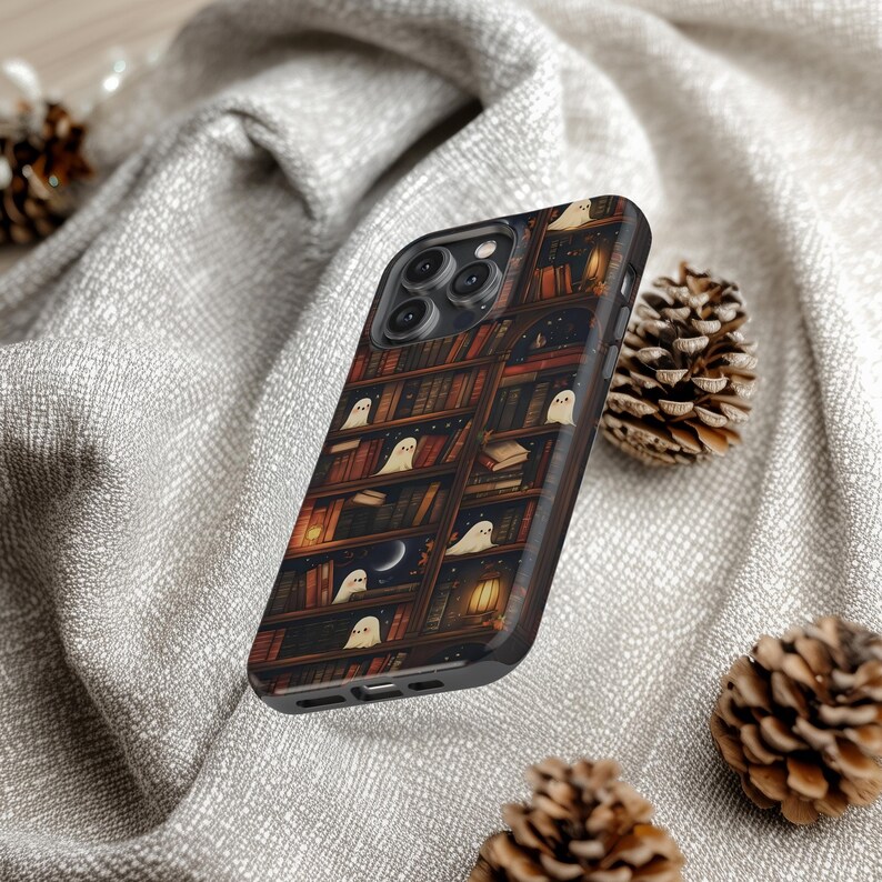 Puede incluir: Una funda negra para smartphone con un dise&ntilde;o caprichoso de una estanter&iacute;a llena de libros y peque&ntilde;os fantasmas amigables. La funda tiene un borde de imitaci&oacute;n de madera oscura y un cielo nocturno con una luna creciente. El tema general es espeluznante y lindo.
