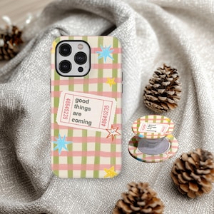 May include: A phone case and matching phone grip with a pink and green plaid design, decorated with blue, yellow, and red stars. The case and grip feature a ticket design with the words "good things are coming".