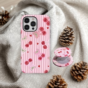 May include: Pink phone case and matching phone grip featuring a cherry pattern with red cherries, green bows, and red hearts on a striped background. The phone case is on a gray surface with pinecones.