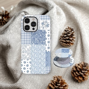 Può includere: Una custodia per telefono e un supporto abbinato con un design patchwork nei toni del blu e del bianco. La custodia è su un tessuto grigio con pigne.
