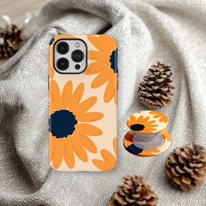 May include: A phone case and phone grip featuring a repeating pattern of orange sunflowers with navy blue centers on a cream background. The phone case is next to a matching phone grip, with pinecones in the background.