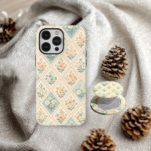 May include: A phone case and phone grip with a floral diamond pattern in cream, light blue, and peach. The phone case is on a gray fabric with pine cones. The phone grip is extended and has a matching design.