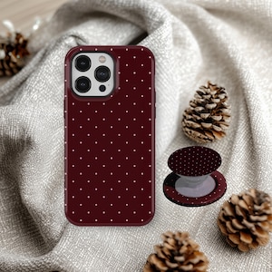May include: A burgundy phone case and matching pop socket with a white polka dot pattern. The phone case is shown next to a pop socket and pine cones on a gray fabric background.