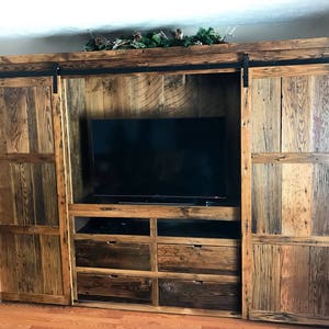 May include: A rustic wooden entertainment unit with sliding barn doors, a large TV space, and multiple drawers. The wood is a mix of brown, grey, and black.