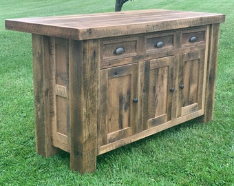 Può includere: Un'isola da cucina in legno rustico con tre ante di armadio e tre cassetti. L'isola ha una finitura in legno invecchiato e ferramenta in metallo nero.