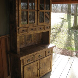 May include: A rustic wooden hutch with glass doors and drawers. The hutch is stained a dark brown and is made from reclaimed wood. It has a distressed look and would be perfect for a farmhouse or country kitchen.