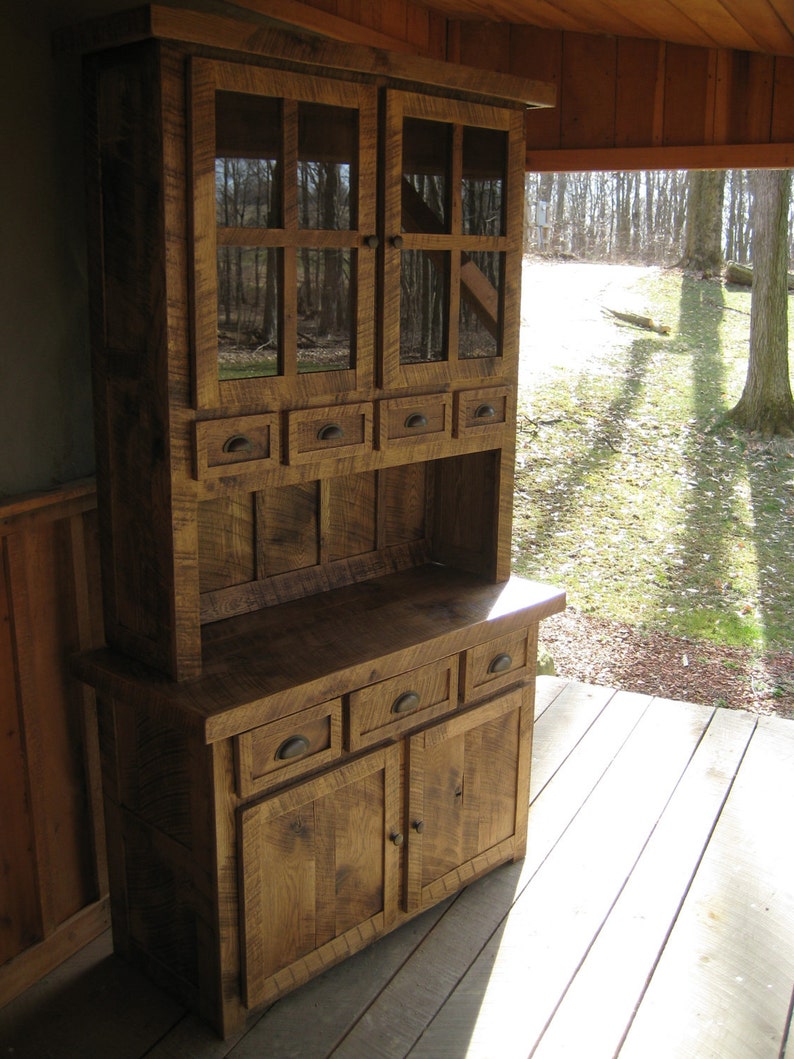 May include: A rustic wooden hutch with glass doors and drawers. The hutch is stained a dark brown and is made from reclaimed wood. It has a distressed look and would be perfect for a farmhouse or country kitchen.
