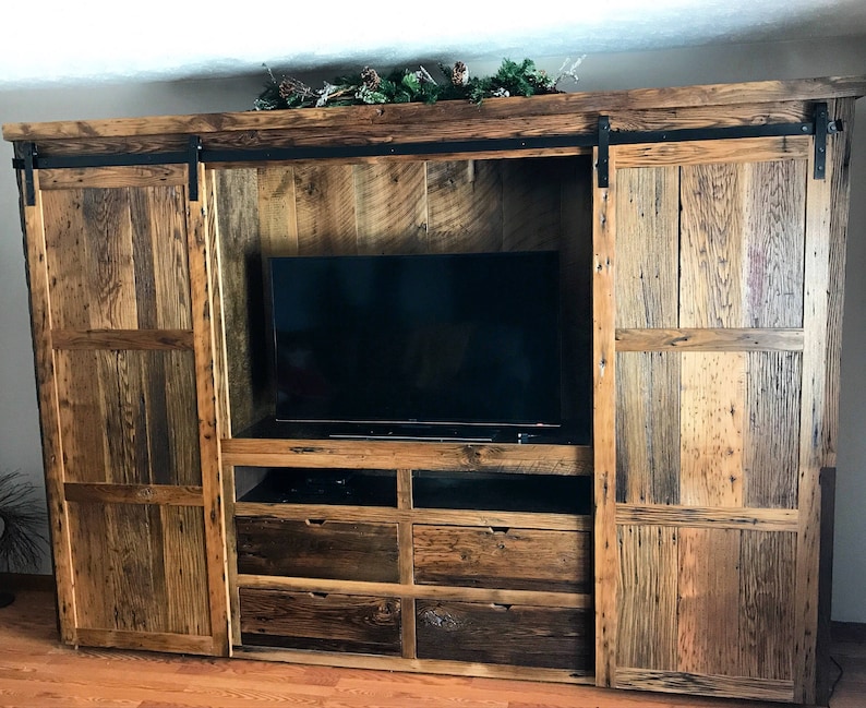 May include: A rustic wooden entertainment unit with sliding barn doors, a large TV space, and multiple drawers. The wood is a mix of brown, grey, and black.