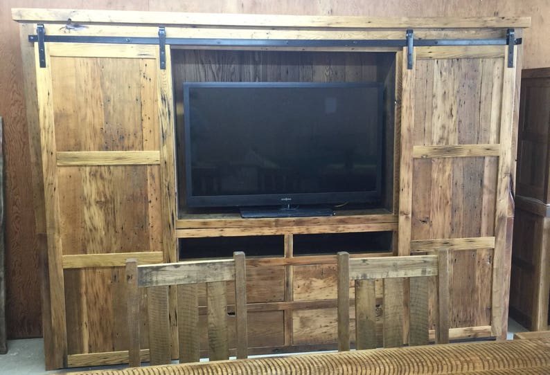 May include: A rustic wooden entertainment unit with sliding barn doors, a flat screen TV, and shelves for storage. The doors are made of reclaimed wood with a natural finish.