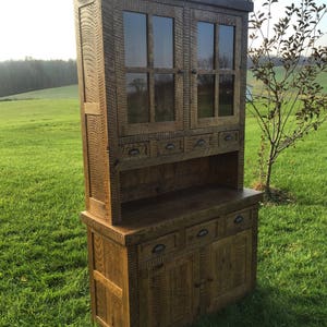 May include: A rustic wooden hutch with glass doors and drawers. The hutch has a distressed finish and is made of reclaimed wood. It has a top section with glass doors and a bottom section with drawers and doors.