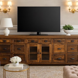 May include: A large, dark wood TV console with multiple drawers and glass-fronted cabinets. A large black television sits on top. The room is decorated with sconces, a white lamp, and a small round table with a bouquet of white flowers.