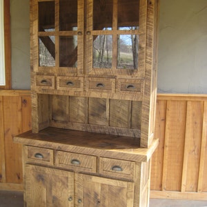 May include: A rustic wooden hutch with glass doors and drawers. The hutch has a distressed finish and is made of reclaimed wood. It features a top cabinet with glass doors and a bottom cabinet with drawers and doors.