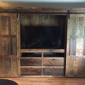 May include: A rustic wooden entertainment unit with sliding barn doors. The unit features a large flat screen TV, four drawers, and open shelves. The doors are made of reclaimed wood with a natural finish.