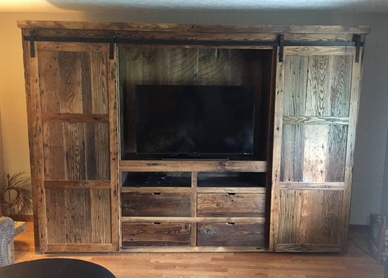 May include: A rustic wooden entertainment unit with sliding barn doors. The unit features a large flat screen TV, four drawers, and open shelves. The doors are made of reclaimed wood with a natural finish.