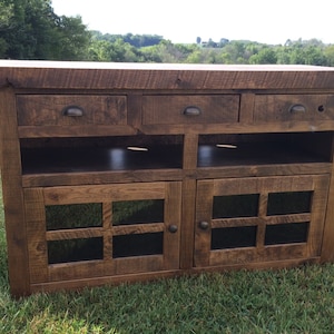 May include: A rustic wooden entertainment center with three drawers, two open shelves, and two glass-paneled doors. The cabinet is made of reclaimed wood and has a dark brown finish.