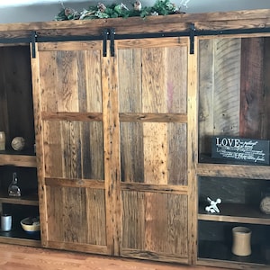 May include: A rustic wooden entertainment unit with sliding barn doors. The doors are made of reclaimed wood and have a natural finish. The unit has shelves for storage and display. The unit is a great way to add a touch of rustic charm to your home.
