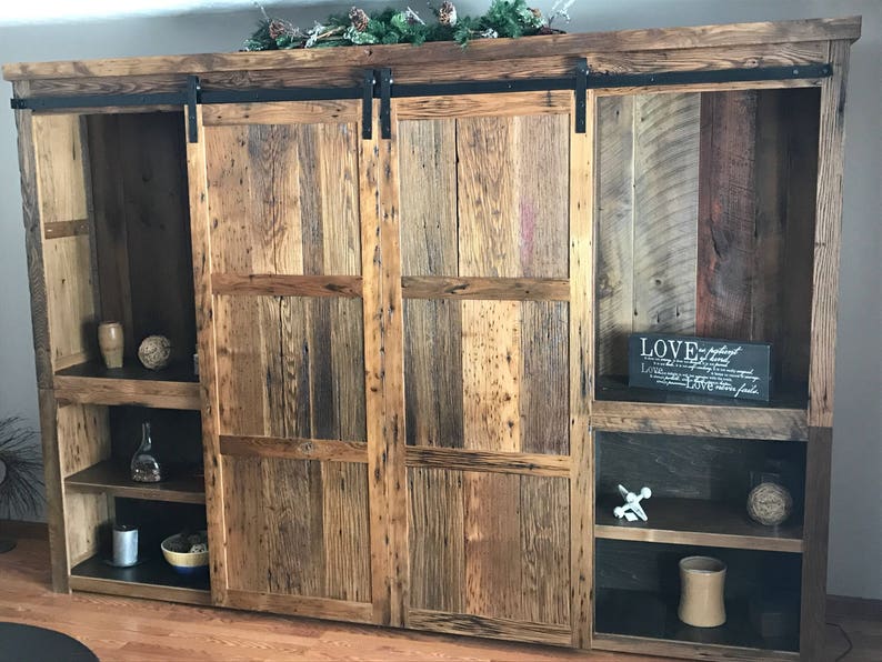 May include: A rustic wooden entertainment unit with sliding barn doors. The doors are made of reclaimed wood and have a natural finish. The unit has shelves for storage and display. The unit is a great way to add a touch of rustic charm to your home.