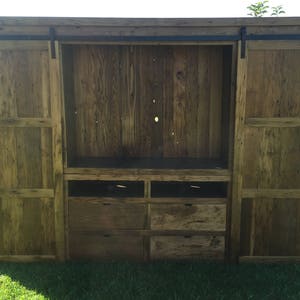 May include: A rustic wooden entertainment unit with sliding barn doors and drawers. The doors are made of reclaimed wood and have a natural finish. The unit has a large open space for a television and shelves for storage.