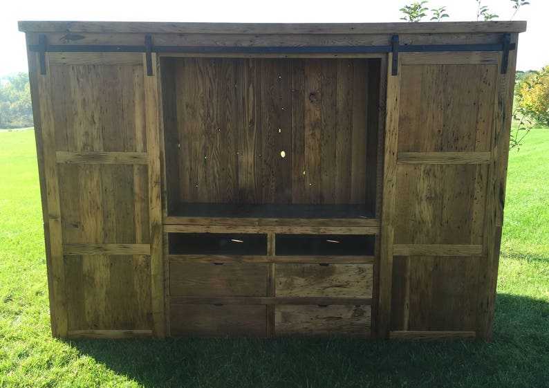 May include: A rustic wooden entertainment unit with sliding barn doors and drawers. The doors are made of reclaimed wood and have a natural finish. The unit has a large open space for a television and shelves for storage.