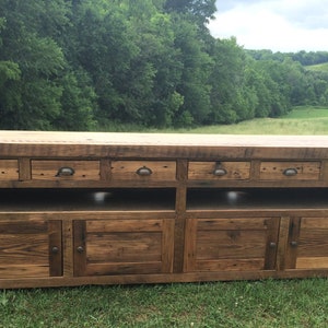 May include: A rustic wooden entertainment center with six drawers, two open shelves, and four cabinet doors. The cabinet is made of reclaimed wood and has a natural finish. The cabinet is sitting on a grassy lawn.