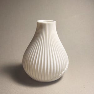 May include: A white, textured vase with a bulbous body and a narrow neck. The vase has vertical, ridged detailing that adds visual interest. The design is simple and modern, suitable for various home decor styles.