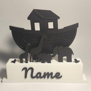 Könnte beinhalten: Ein schwarzer Tortenaufsatz mit der Arche Noah, einem Boot, Tieren und dem Wort "Name". Die Arche hat eine hausartige Struktur und Herz-Ausschnitte. Zu den Tieren gehören Giraffen, Elefanten und Vögel. Die Basis ist weiß.