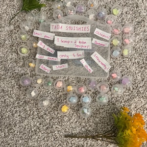 May include: A display of Taba Squishies, each in a clear capsule, containing small, colourful toys. Handwritten labels are visible, with phrases such as "small paw!" and "Strawberry!" and a price.