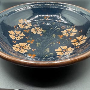 May include: A round ceramic bowl with a dark blue interior and a brown rim. The bowl is decorated with a floral pattern in shades of tan and orange. The flowers are arranged around a central stem.