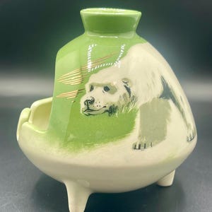 May include: A ceramic ashtray with a polar bear design. The ashtray is primarily white and green, with a three-legged base. The polar bear is depicted in a realistic style, with black and gray details.