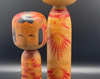 May include: Two wooden Kokeshi dolls. The dolls have a light brown base color with red and black painted details. The larger doll has a cylindrical body with a floral design. The smaller doll has a similar design. Both dolls have a rounded head with painted facial features.
