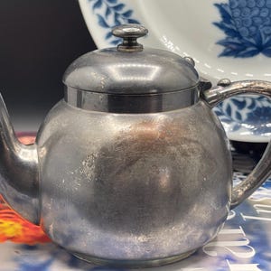 May include: A small, silver-colored teapot with a rounded body, spout, and handle. The lid has a small knob on top. A white plate with blue floral designs is in the background. The teapot appears to be made of metal and has a vintage look.