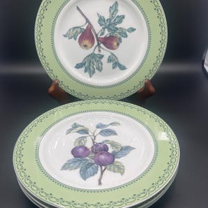 May include: A collection of white ceramic plates with a pale green border and fruit illustrations. One plate shows figs, others feature plums. The plates are stacked and displayed on a dark surface.
