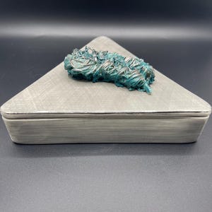 May include: A gray triangular box with a silver trim and a teal-colored, textured, decorative element on top. The box has a lid and a rectangular base. The background is black.