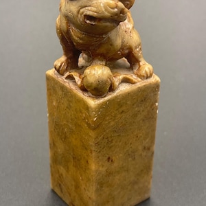 Può includere: Scultura in pietra marrone chiaro raffigurante una creatura mitica, forse un leone guardiano, su una base rettangolare. La scultura presenta dettagli, tra cui un'espressione accigliata. La base è quadrata. Lo stile suggerisce un oggetto antico o vintage.